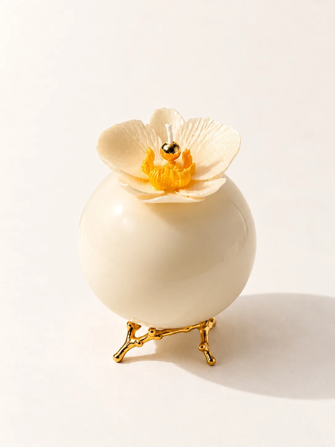 Vela esférica cor de marfim com uma flor no topo, sobre suporte dourado